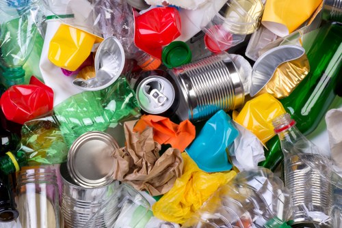 A close-up view of a collection of mixed recyclable waste materials scattered on a surface, including crushed and uncrushed plastic bottles in various colours such as green, clear, and yellow, with some featuring labels and caps. There are several empty aluminium cans, one prominently positioned in the centre with a partially opened pull tab, displaying their smooth metallic surfaces, alongside crumpled brown paper and plastic bags in shades of orange, blue, and white. The background appears to be a clean, light-colored surface, possibly outdoors or in a waste collection area, with some items overlapping each other. This image illustrates typical waste seen in rubbish removal services, such as those offered by Commercial Waste Shepperton, conveying the importance of proper disposal and recycling of household and commercial waste in the Shepperton area, within the postal code region related to TW or KT postcode districts.