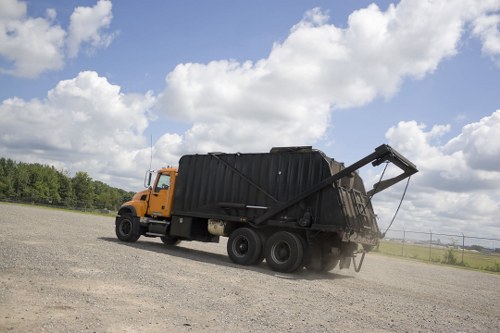 A large yellow and black waste collection truck parked on a flat, gravelly surface under a partly cloudy sky. The truck has a sturdy metal body with visible texture, featuring a black rear compactor section with an open top and sloped sides. The vehicle's cab is painted yellow, with black details on the grille and wheels. The compactor section appears to be used for transporting bulky rubbish, with a hydraulic arm mechanism partially visible at the front. Surrounding the scene are open fields with sparse vegetation in the distance, and the sky shows scattered white clouds against a blue backdrop. This image likely relates to rubbish removal services offered by Commercial Waste Shepperton, serving the local area with waste disposal solutions for commercial clients.