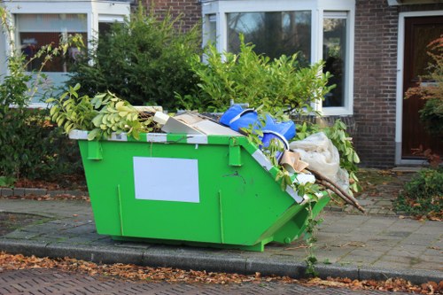 A large, bright green rectangular skip positioned on a paved area in front of a residential building. The skip is filled with various types of garden and household waste, including green leafy branches, plastic bags, cardboard boxes, and other mixed rubbish. The waste extends above the rim of the skip, with some items slightly leaning or spilling over the edges. The skip's surface appears to be made of metal with a clean finish, and it is situated directly on the pavement, bordered by a curb. Behind the skip, there is a brick house with white window frames, a bay window, and a sliding door, surrounded by bushes and small trees, indicating a suburban setting. The scene is well-lit with natural daylight, emphasizing the contrast between the vivid green of the skip and the mixed array of waste materials, illustrating a typical rubbish collection situation for a property in Shepperton, served by Commercial Waste Shepperton.