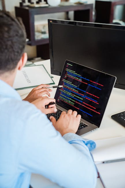 A person working at a desk, viewed from behind, with short dark hair and wearing a light blue shirt. They are using a laptop that displays lines of colorful computer code on a dark background. The des